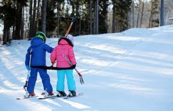 Zwei Kinder stehen auf ihren Skiern, verbunden durch ein Seil, während sie den sanften Hang hinaufgezogen werden. Die strahlende Wintersonne lässt den Schnee glitzern und schafft eine fröhliche Atmosphäre, die die Freude am Skifahren in der Familie widerspiegelt.