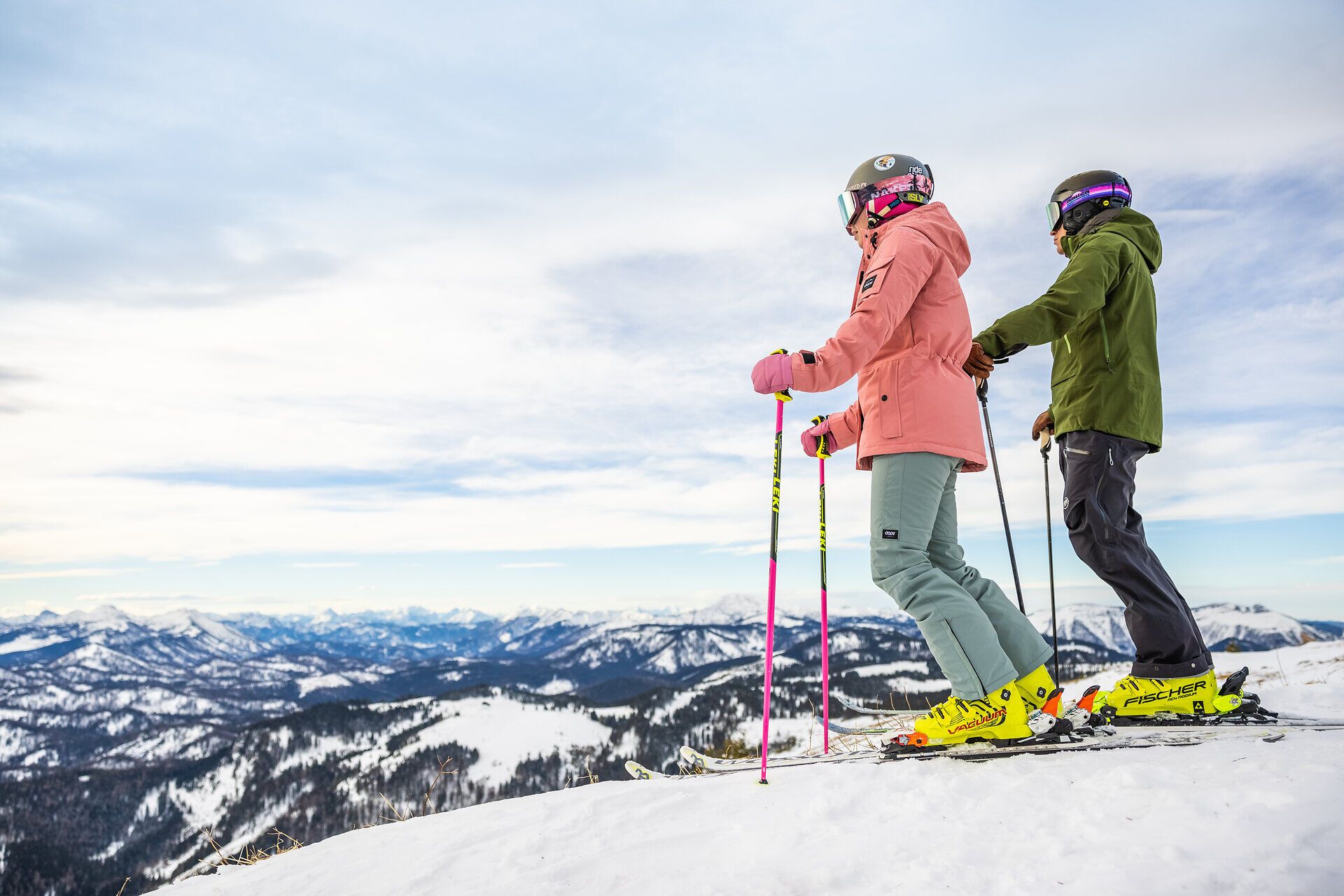 Ein Paar genießt die frische Bergluft und die atemberaubende Aussicht auf die schneebedeckten Gipfel, während sie sich auf das Skifahren vorbereiten. Die sanften Hügel und die klare Winterluft schaffen eine perfekte Kulisse für unvergessliche Momente im Schnee.