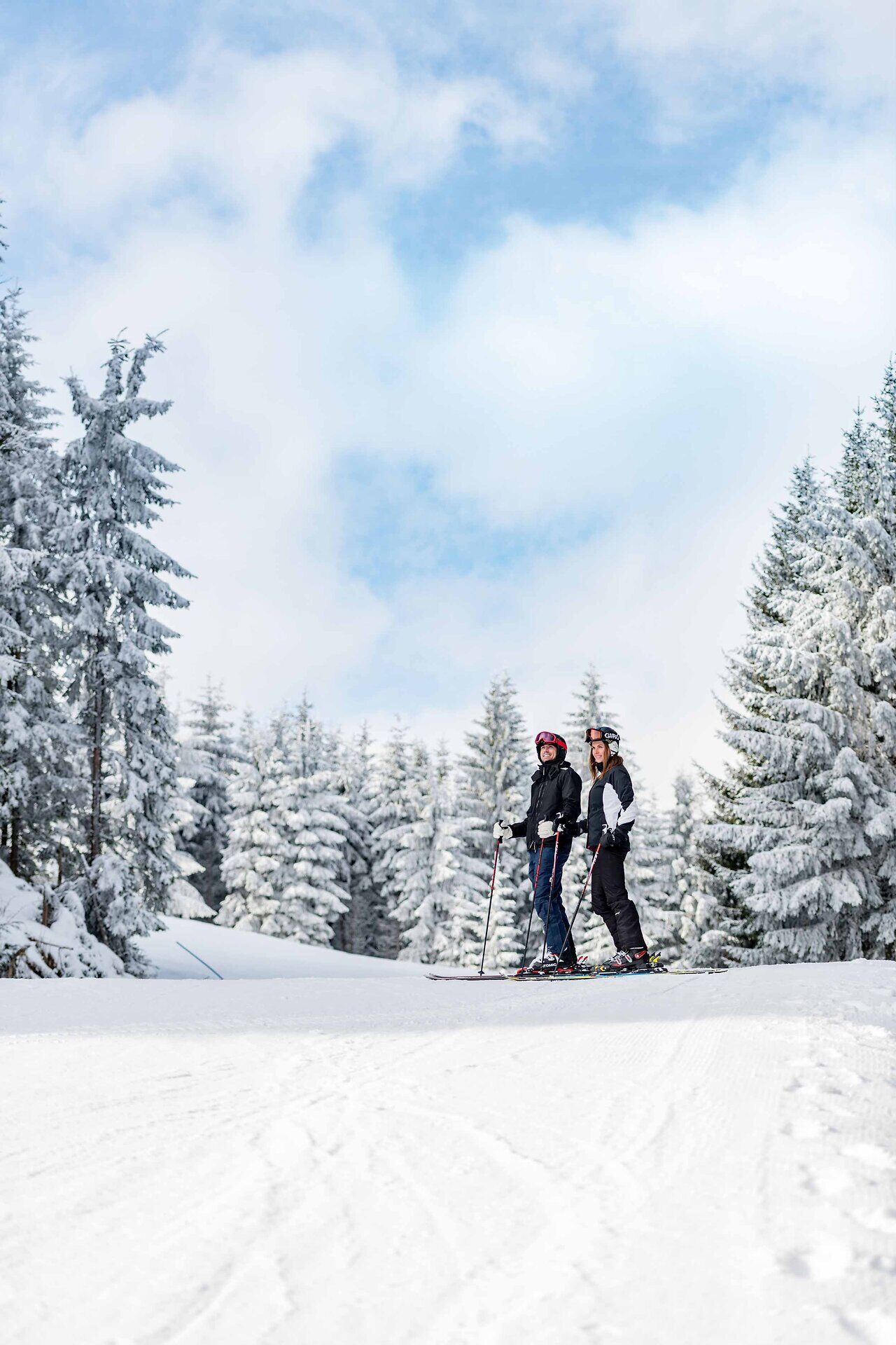 Inmitten einer traumhaften Winterlandschaft stehen zwei Skifahrer bereit, die frische Bergluft einzuatmen. Die schneebedeckten Bäume um sie herum funkeln im Sonnenlicht und schaffen eine magische Atmosphäre, die zum Skifahren einlädt.