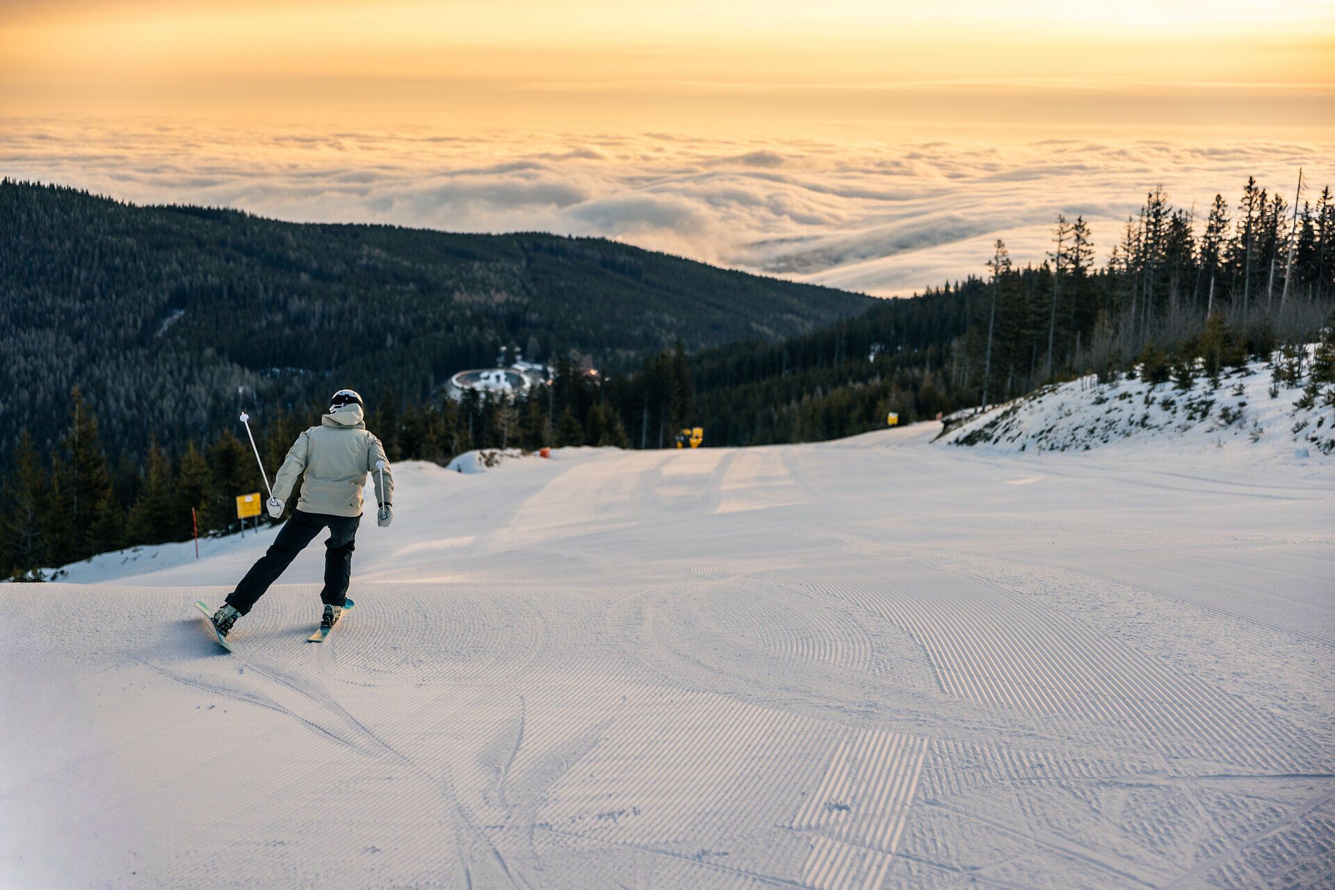Skifahrende Person im Vordergrund auf einer präparierten Skipiste mit Blick über verschneite Berghänge und Wolken bei tief stehender Sonne.