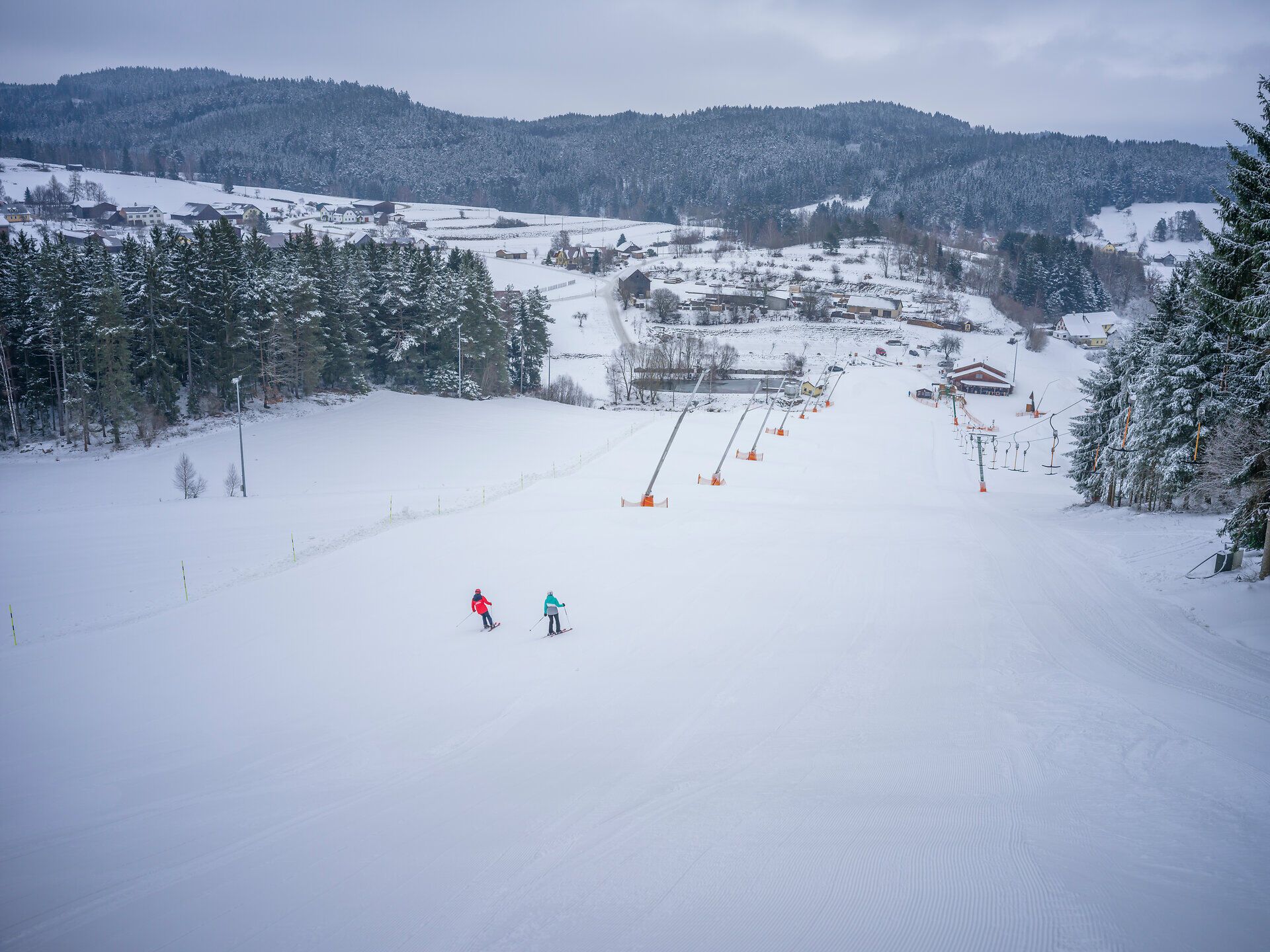 Die sanften, schneebedeckten Hänge laden zu einem unvergesslichen Skierlebnis ein. Zwei Skifahrer gleiten harmonisch über die frisch präparierte Piste, während die winterliche Landschaft in ihrer vollen Pracht erstrahlt. Hier, umgeben von majestätischen Tannen und der ruhigen Atmosphäre der Berge, wird der Winter zum wahren Erlebnis.