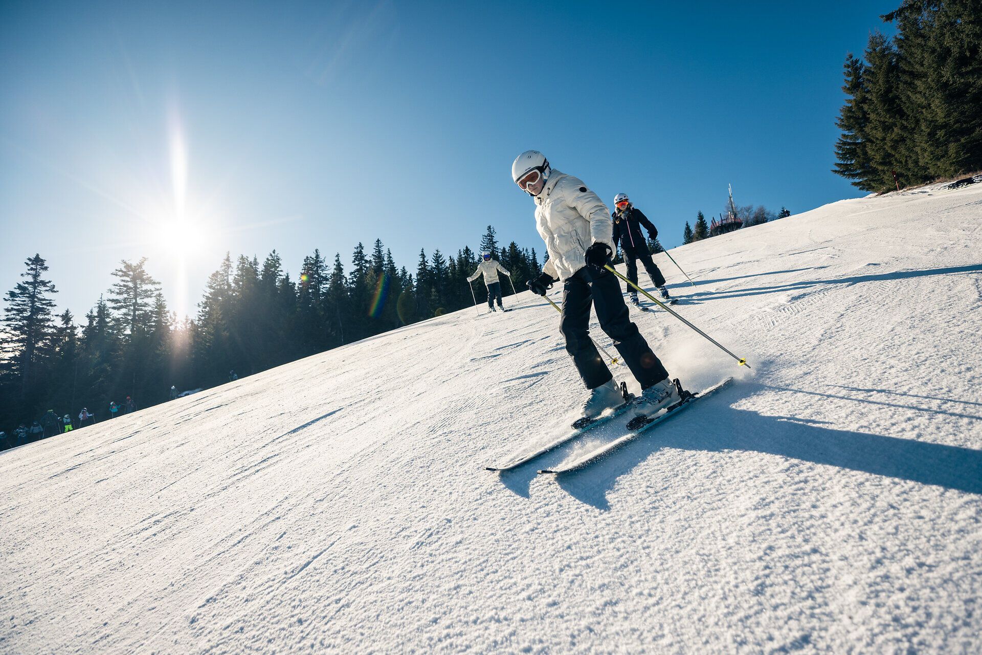 Mehrere Personen mit Skiern auf einer verschneiten Skipiste der Erlebnisalm Mönichkirchen, umgeben von Nadelbäumen und winterlicher Berglandschaft.