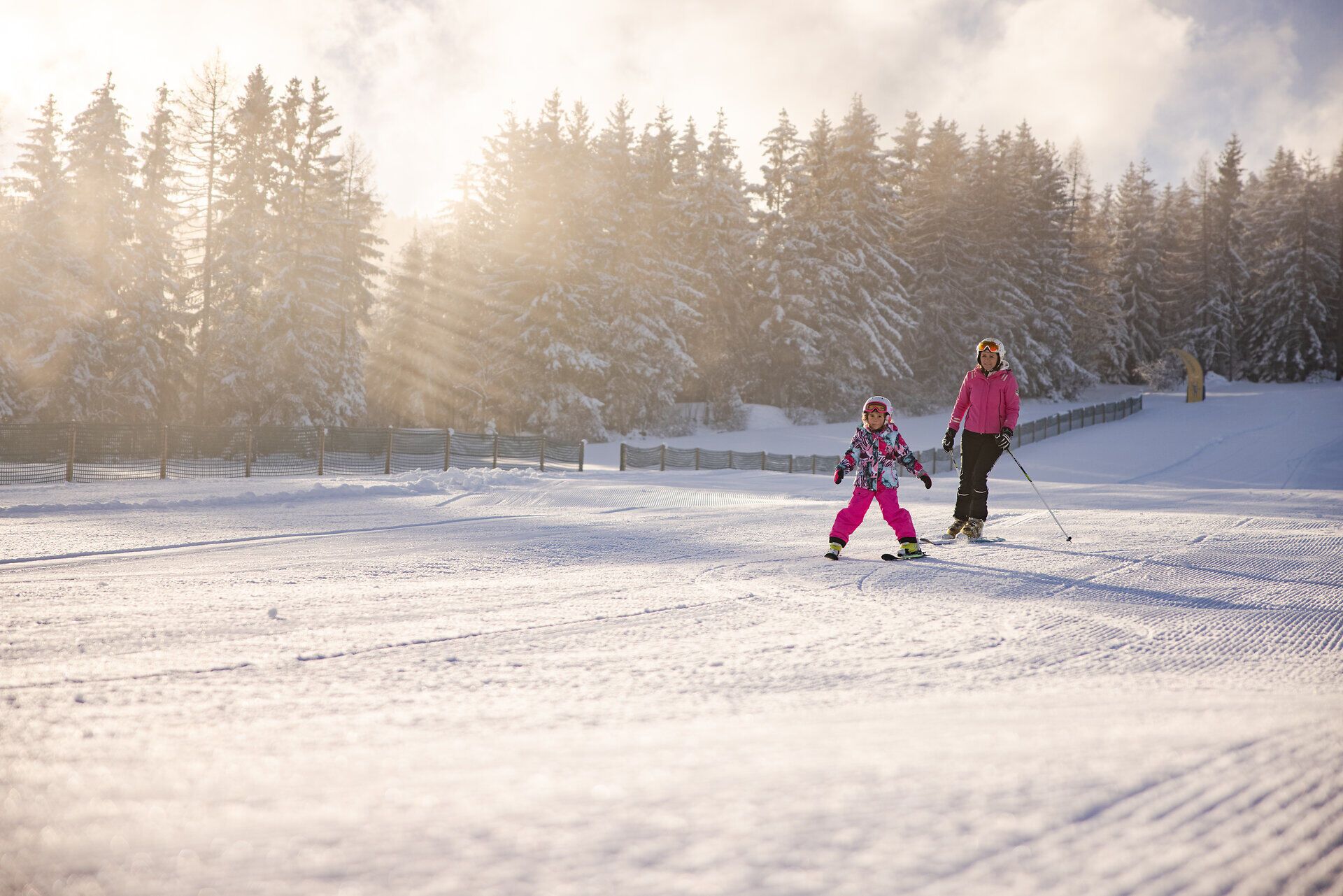 Kind beim Skifahren auf einer sonnigen Skipiste, begleitet von einer erwachsenen Person